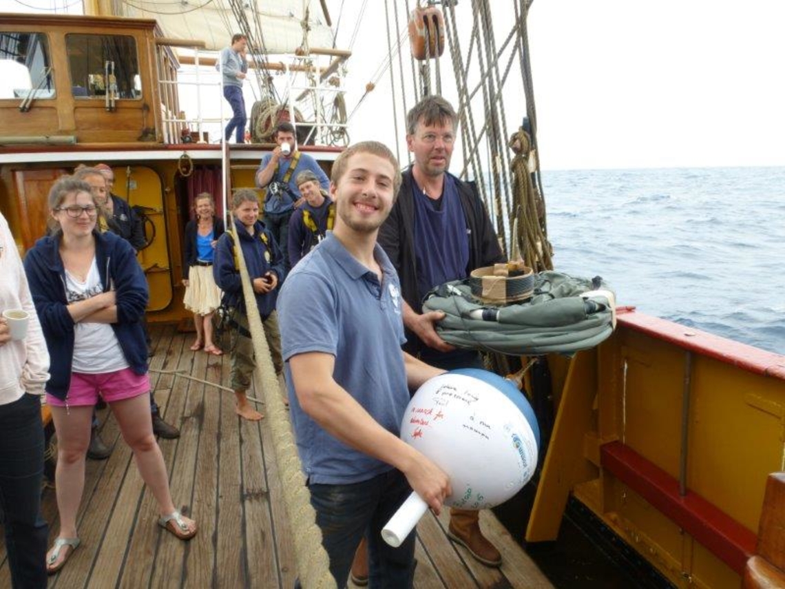 A smiling scientist is holding a drifting buoy ready to be deployed. Another is slightly behind, holding the drogue. They are both onboard a ship.